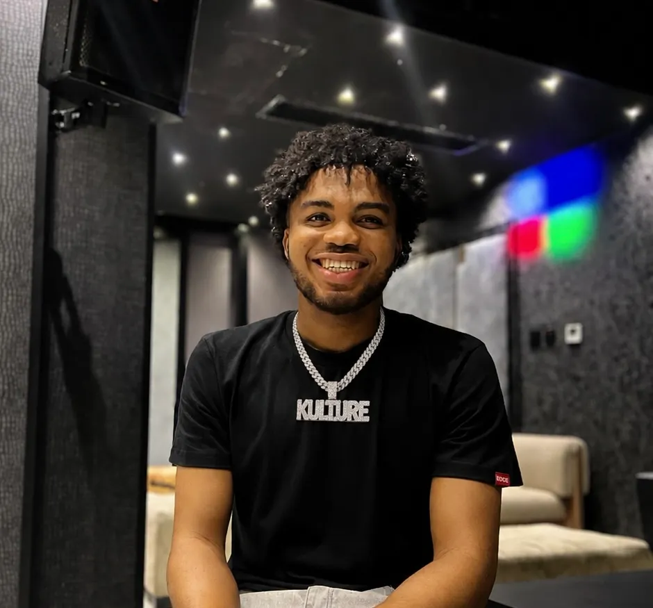 Kul-Ture smiling in a black shirt and chain in a dim, colorful studio