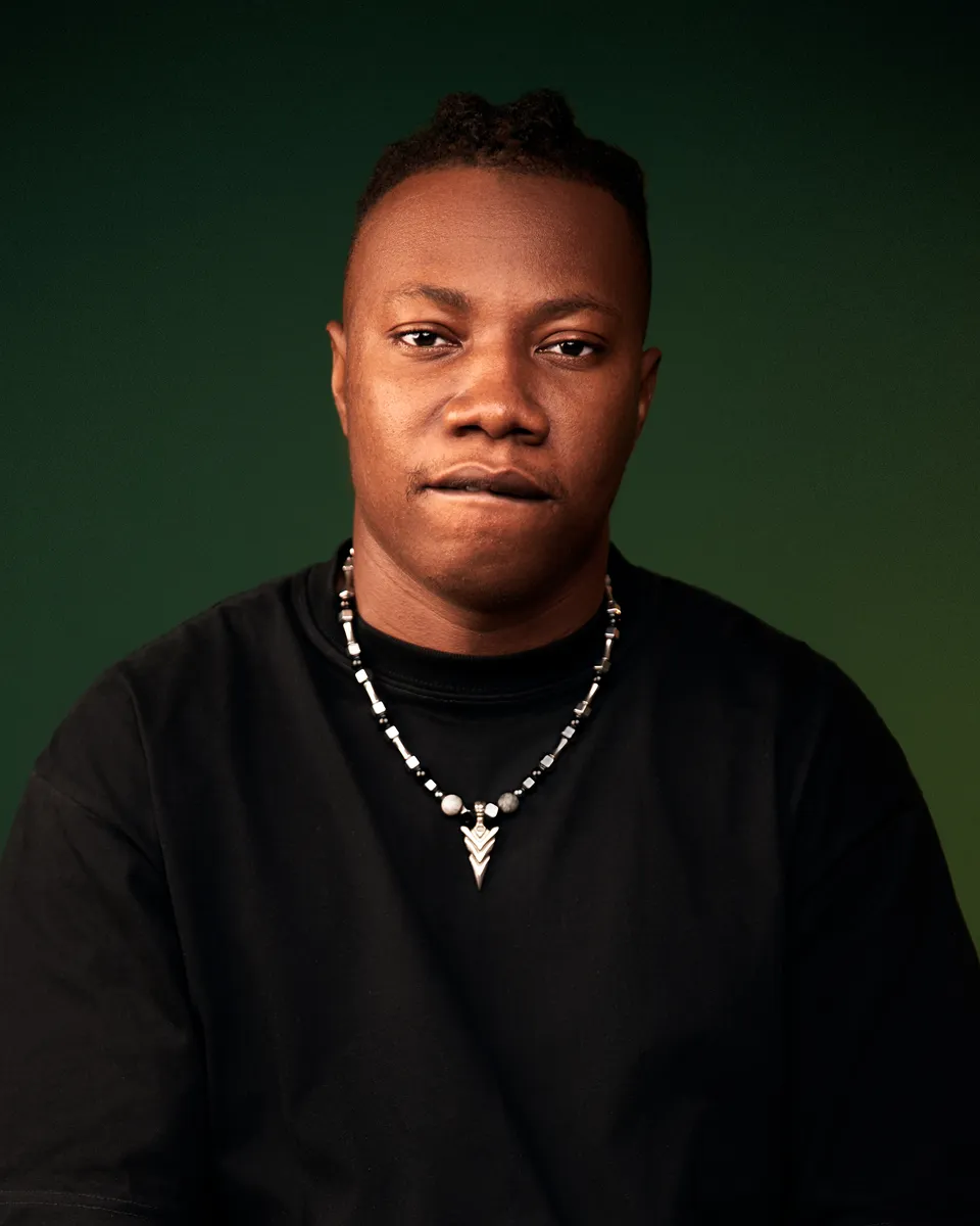 Lyl Bobby in a black shirt and necklace against a dark green backdrop