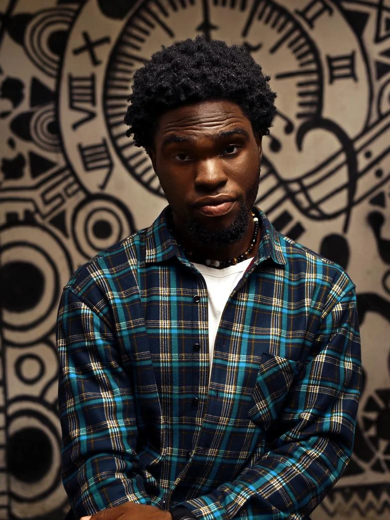 DJ MRGN posing in a plaid shirt against a patterned wall