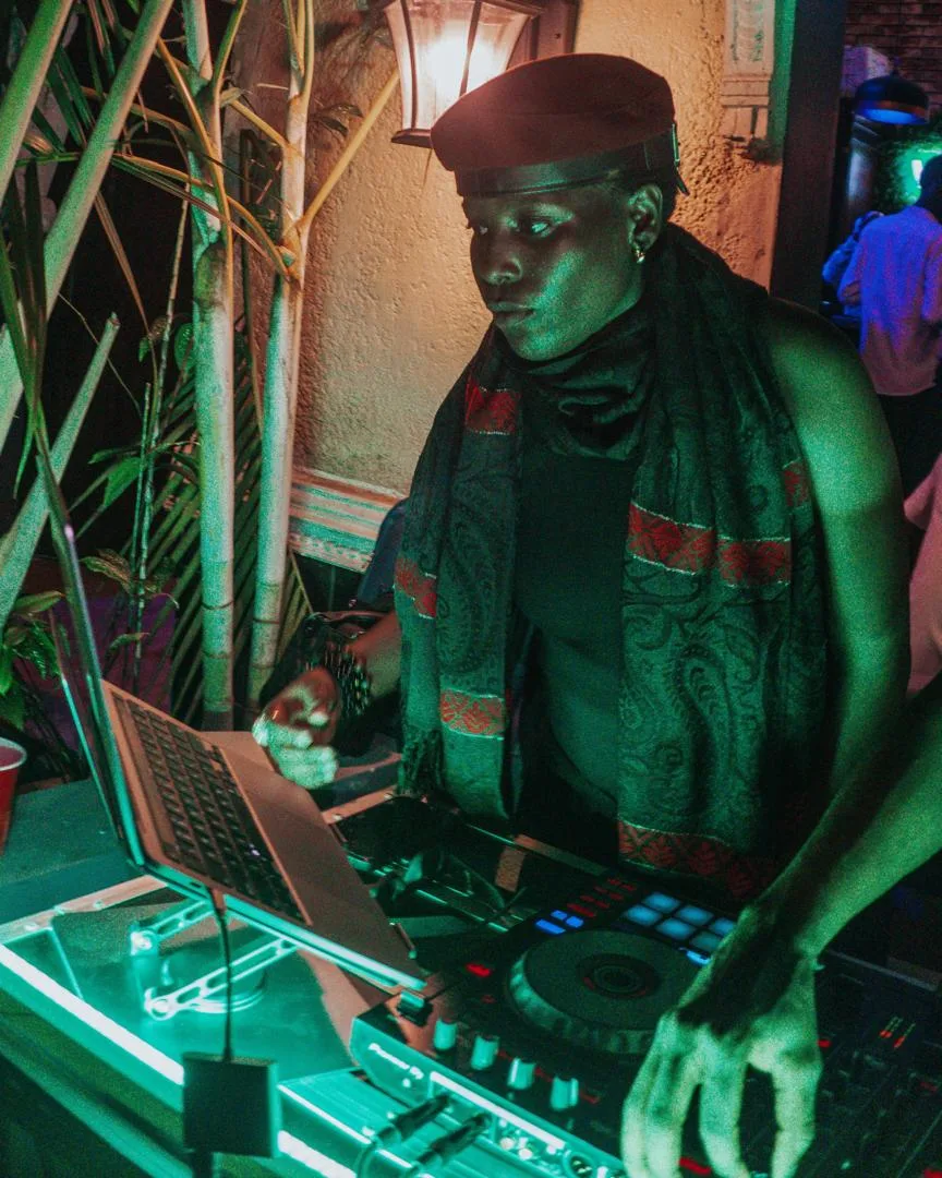 DJ Nacci in a cap and scarf performing at a mixing console under green and red lights