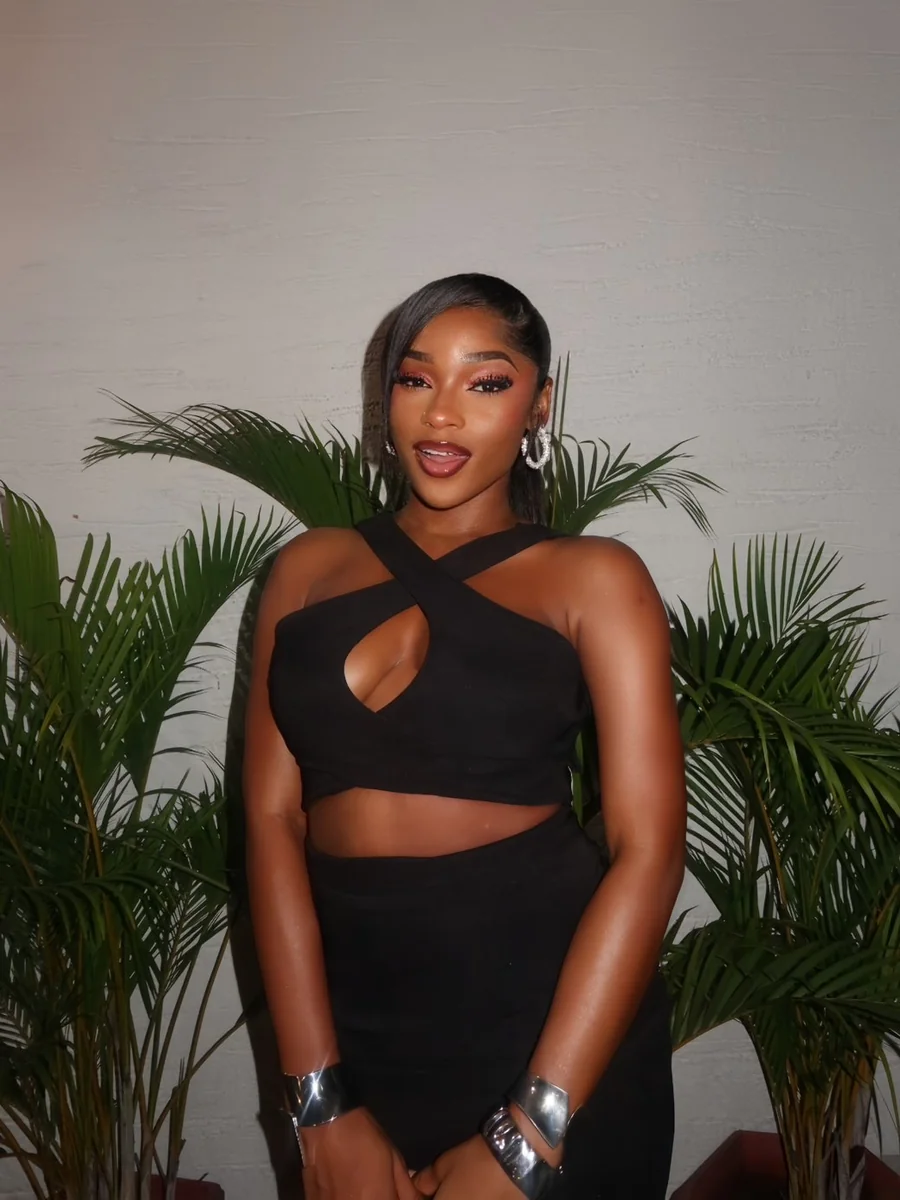 DJ TraeWhay posing in a black outfit surrounded by palm leaves