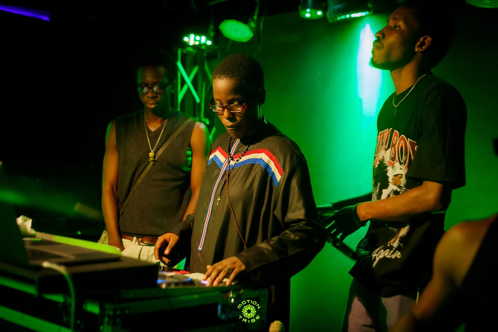 Two performers stand beside a DJ controller under vivid green stage lighting
