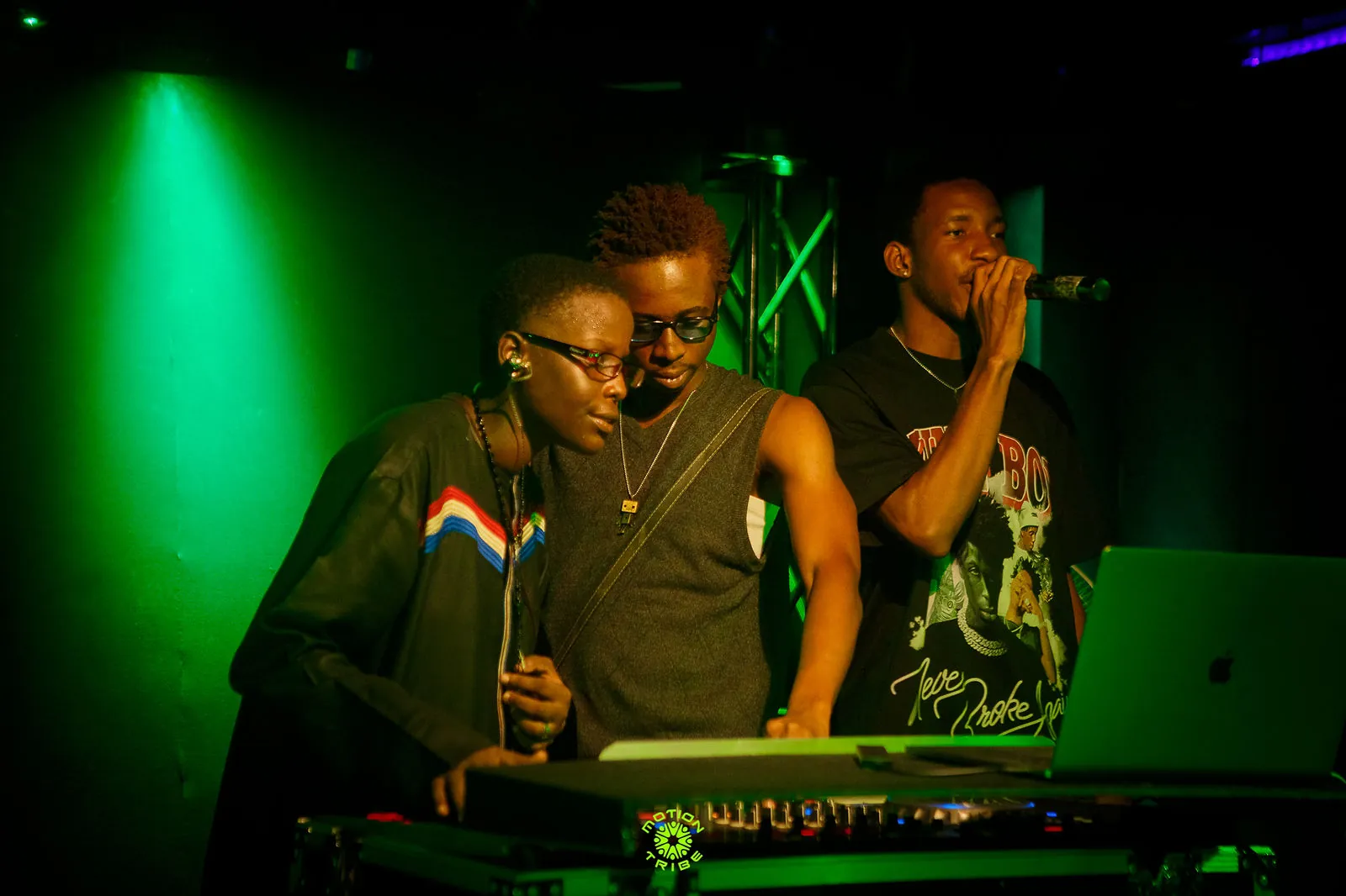 Three performers gather around a DJ setup, with one holding a microphone